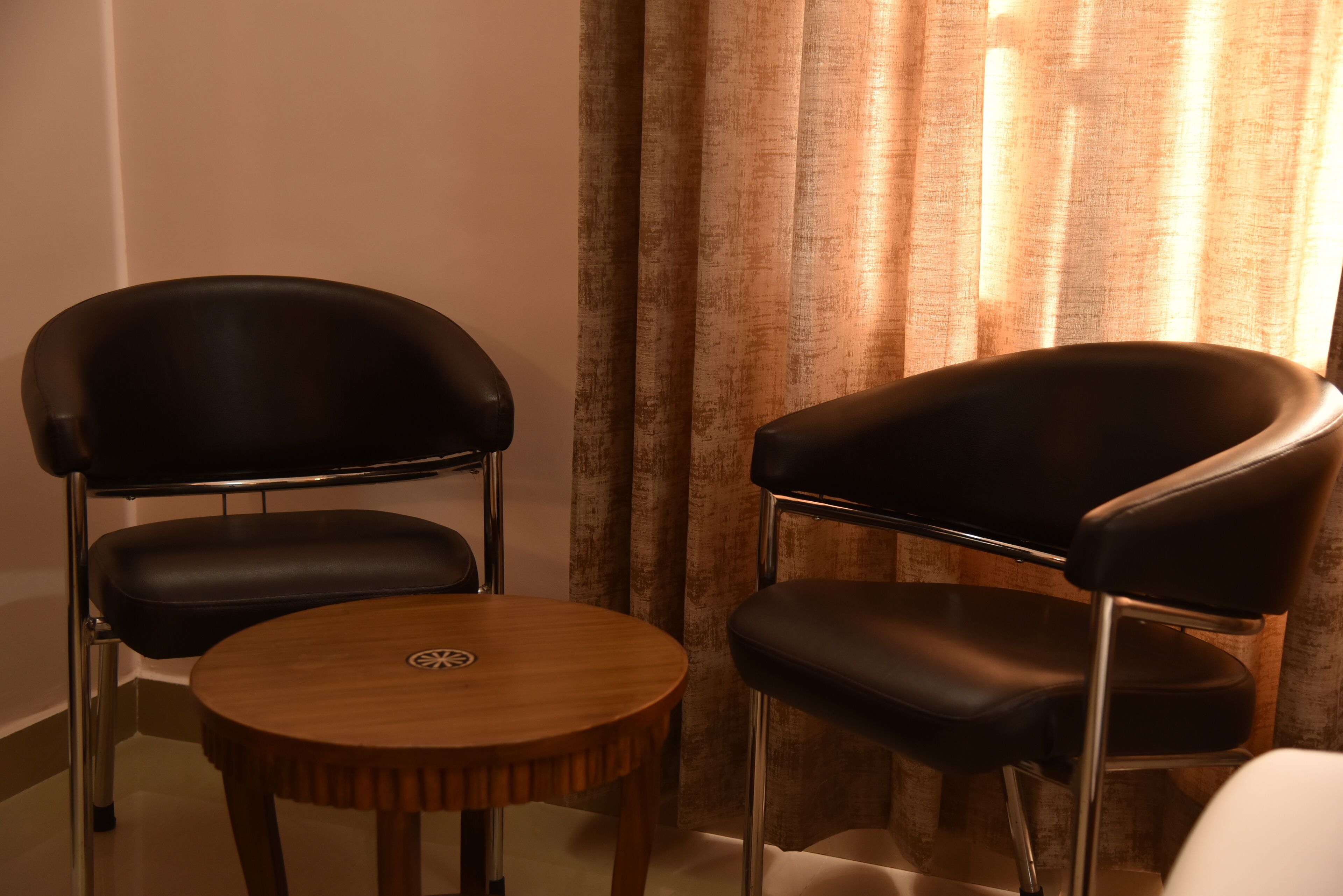 Two black leather chairs and a round wooden table in a room with a curtain in the background.