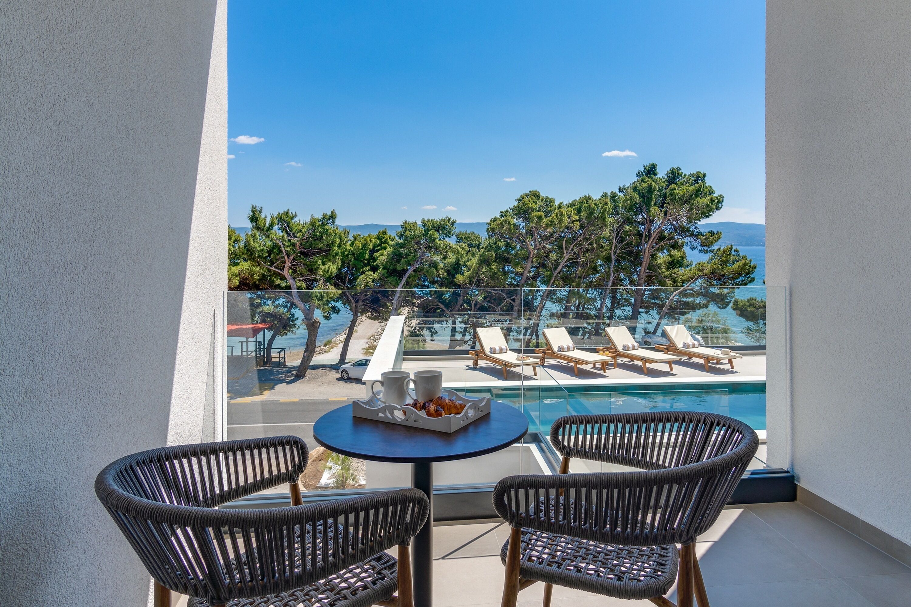 A balcony with wicker chairs, a table with a tray of food, and a view of a pool and lounge chairs.
