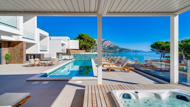 Un espace piscine extérieur moderne, doté de chaises longues et d’un jacuzzi, offrant une vue imprenable sur une mer d’un bleu profond et sur une montagne.