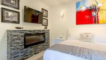 A bedroom with a stone fireplace, a large bed, a TV mounted on the wall, and framed pictures.