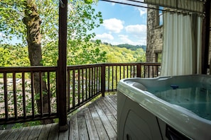 A wooden deck with a hot tub, a view of a forested landscape, and a stone building.