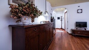 A hallway with wooden flooring, a dark wooden cabinet with drawers, a white vase with dried flowers, a hanging pendant light, a clock on the wall, and a television on a wooden stand. - Italianway - Gradisca 8 (Udine)