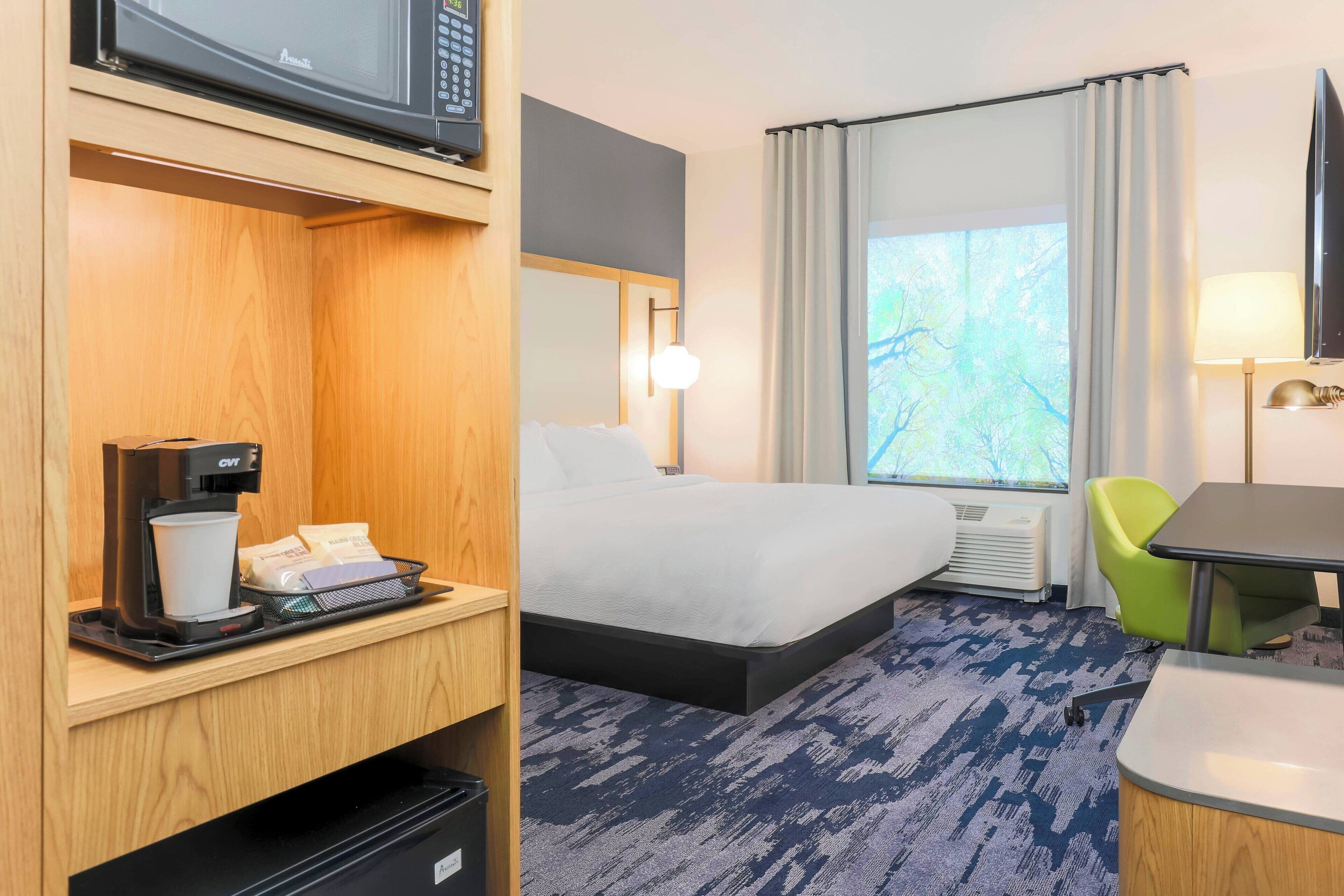 A hotel room with a bed, a desk with a coffee maker, a chair, a TV, and a window with curtains.