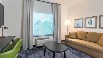 Ein Hotelzimmer mit Sofa, kleinem Tisch, grünem Sessel, Flachbildfernseher, Fenster mit Vorhängen, Wandleuchte und gerahmten Kunstwerken.