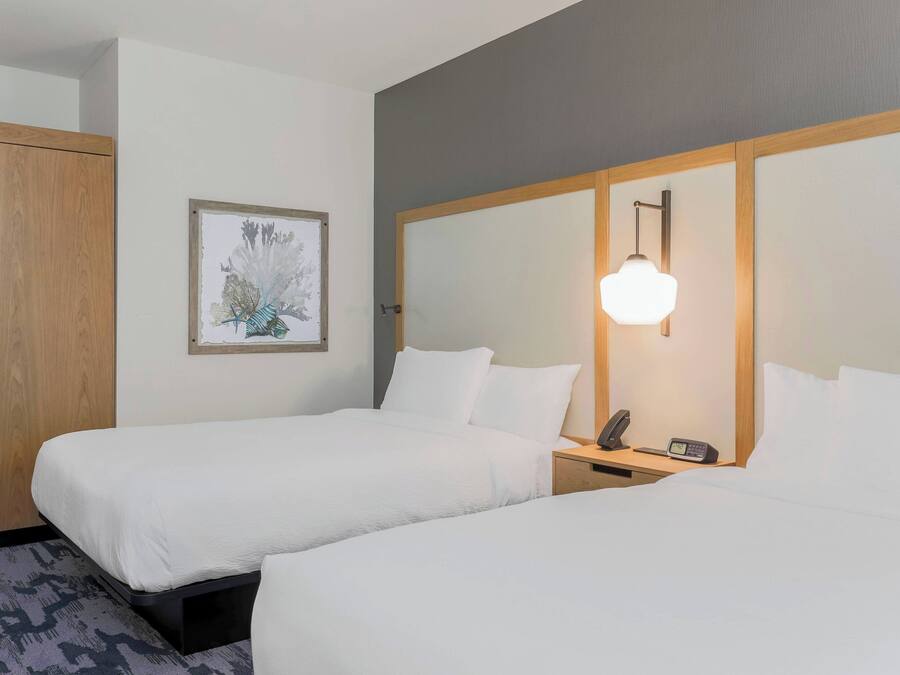A hotel room with two beds, a wooden wardrobe, a framed picture on the wall, and a bedside table with a lamp and phone.