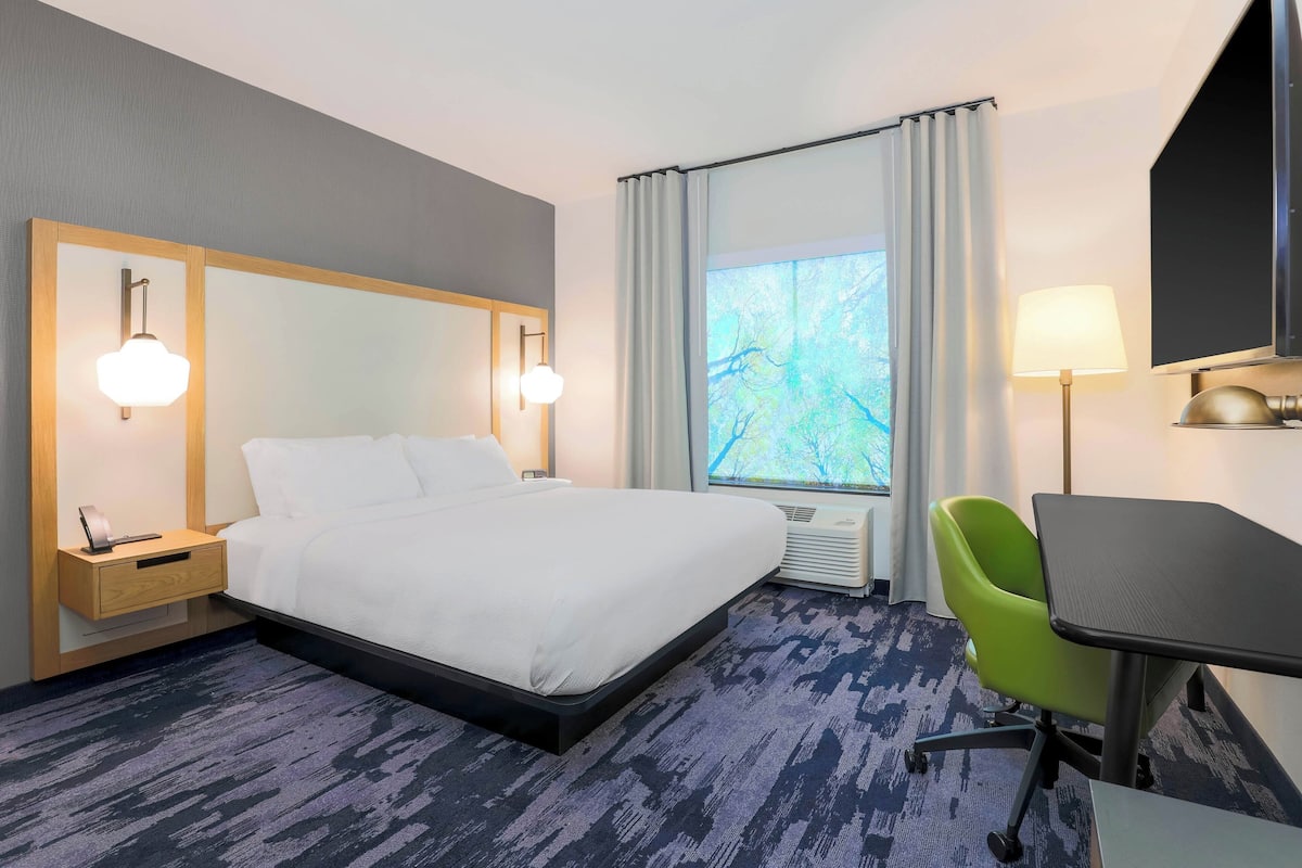 A hotel room with a bed, bedside table, lamp, chair, desk, and window with curtains.