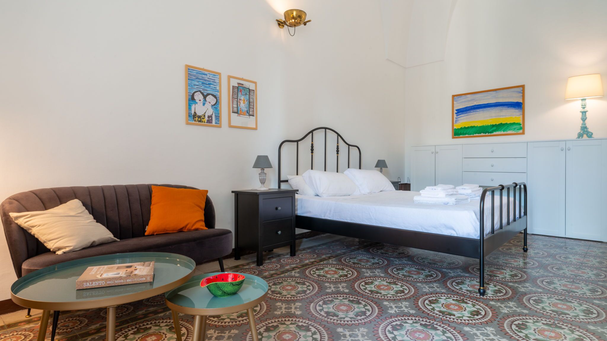 Een slaapkamer met een bed, een slaapbank, een nachtkastje, een lamp, een klein tafeltje met een boek en een vloer met een patroon.