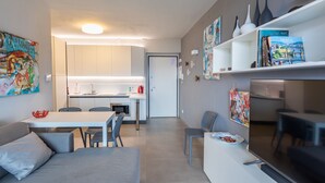 A modern living room with a sofa, a dining table with chairs, a kitchen area with a fridge, and a TV stand with a TV.