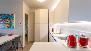 A modern kitchen with white cabinets, a white countertop, and red kitchen appliances. There is a dining area with a white table and chairs, and a colorful painting on the wall.