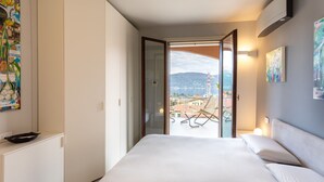 A bedroom with a bed, a view of a balcony with chairs, and a cityscape in the background.