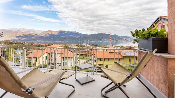A balcony with two wicker chairs and a small table, overlooking a town with buildings and mountains in the background.