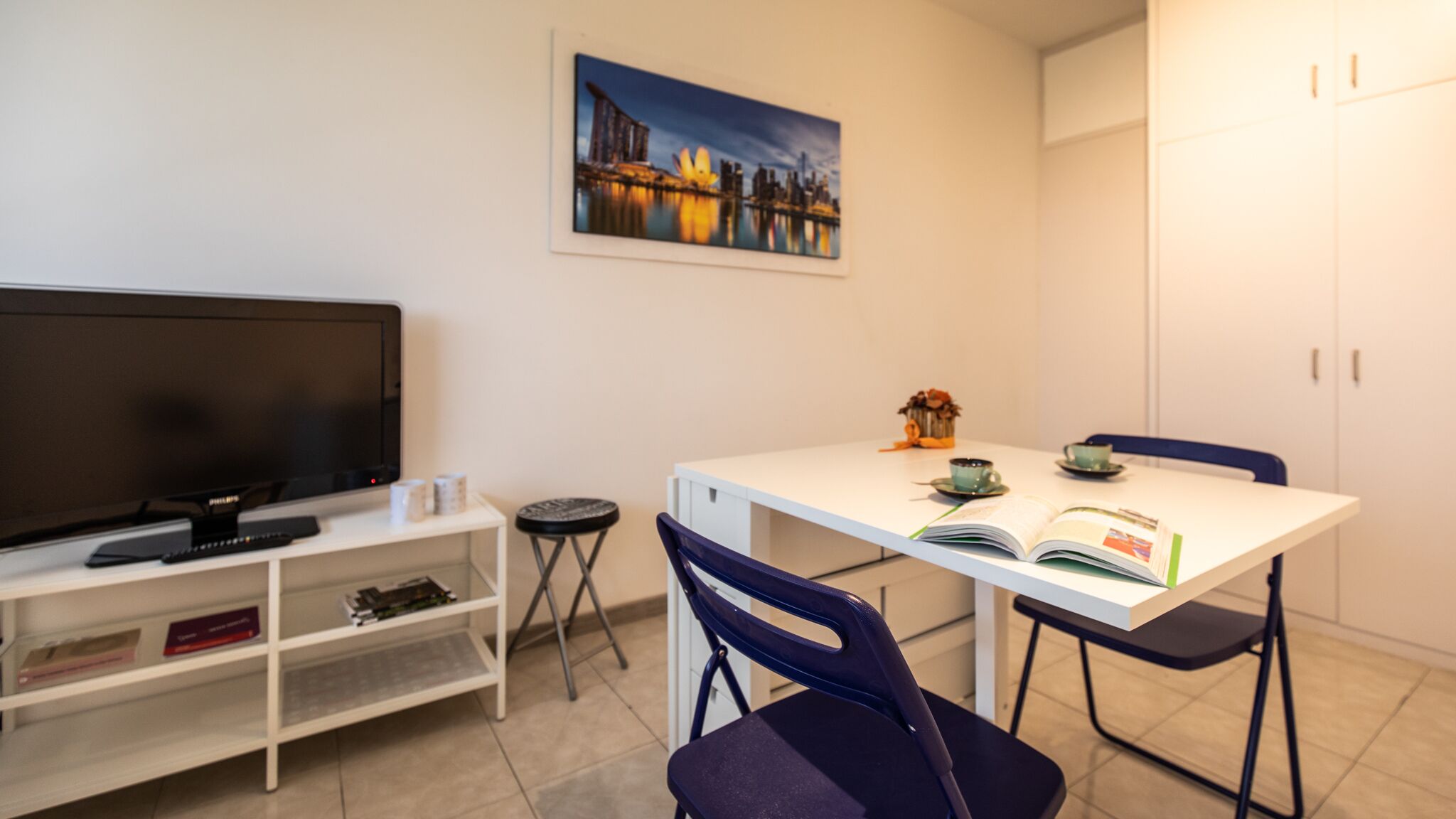 Een compacte, moderne kamer met een witte tafel, twee stoelen, een televisie en een schilderij aan de muur.