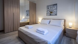 A neatly made bed with folded white towels, a wooden wardrobe, a bedside table with a lamp, and a large flower painting on the wall. - Italianway  - Bersaglio 25 (Udine)