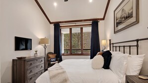 A bedroom with a bed, a dresser, a nightstand, a lamp, a television, and a window with curtains.