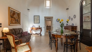 A dining area with a round table set for six, surrounded by chairs. There is a sofa and a lamp in the foreground. - Italianway - Casa del Barone (Lecce)