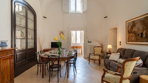 A dining area with a round table, chairs, and a vase of flowers. There is a television on a stand and a painting on the wall. - Italianway - Casa del Barone (Lecce)