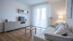 Een moderne woonkamer met een flatscreen televisie, een houten salontafel, een bank en een groot raam met dunne gordijnen.