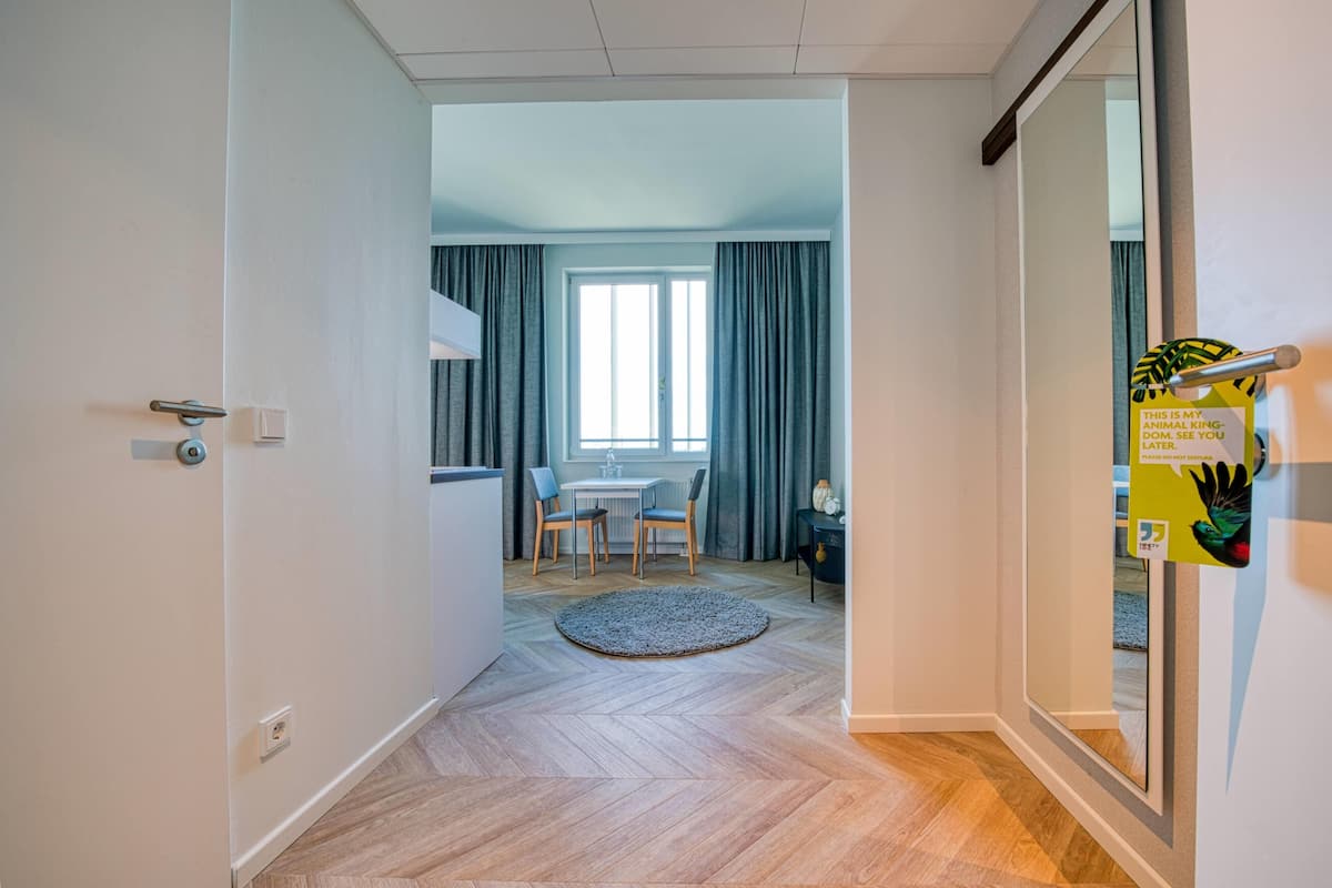 Een moderne hotelkamer met een houten vloer, een rond tapijt, een zithoek met een tafel en stoelen, en een groot raam met gordijnen.