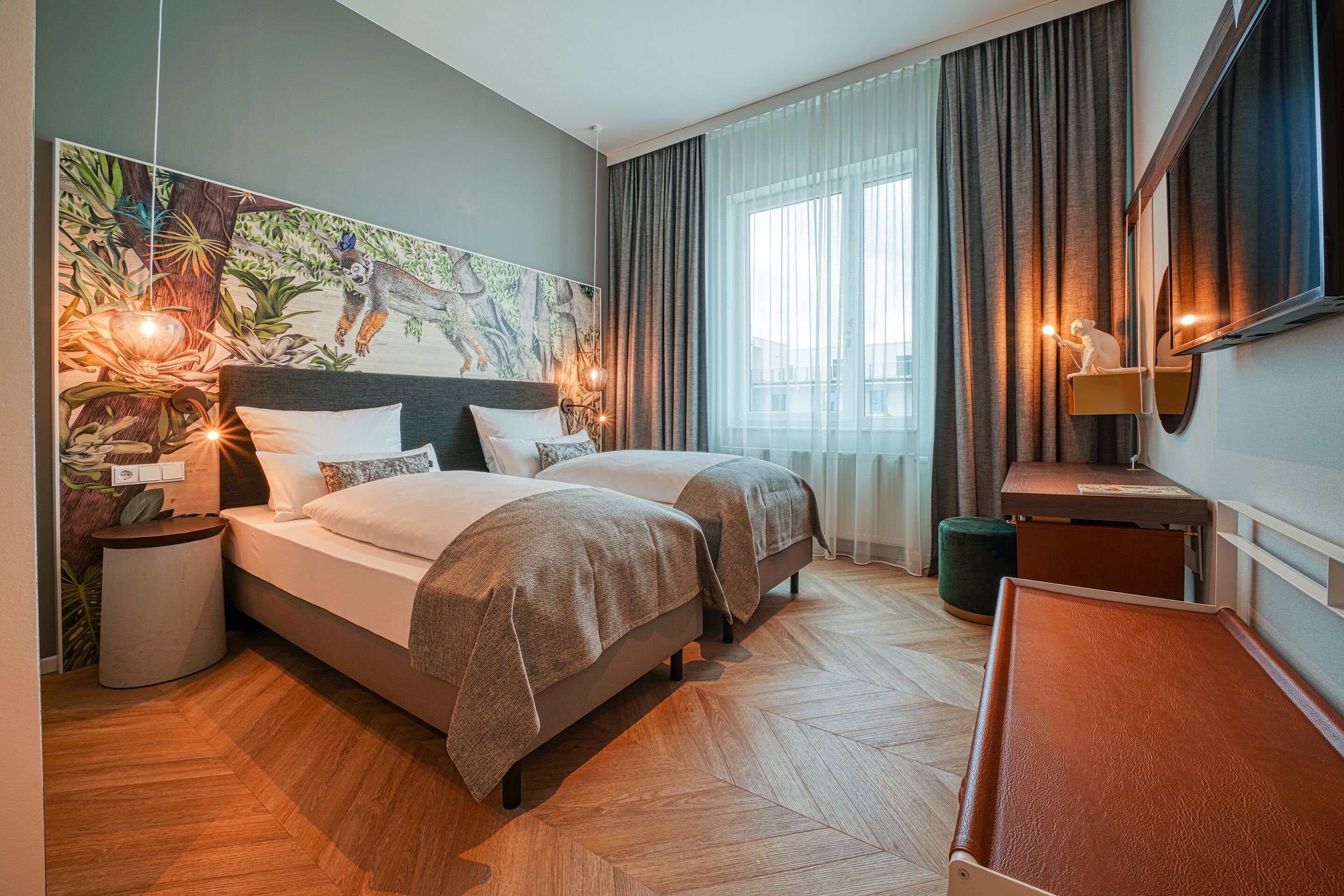 A hotel room with two beds, a large mural on the wall, a desk with a mirror, and a view of the outdoors through large windows.