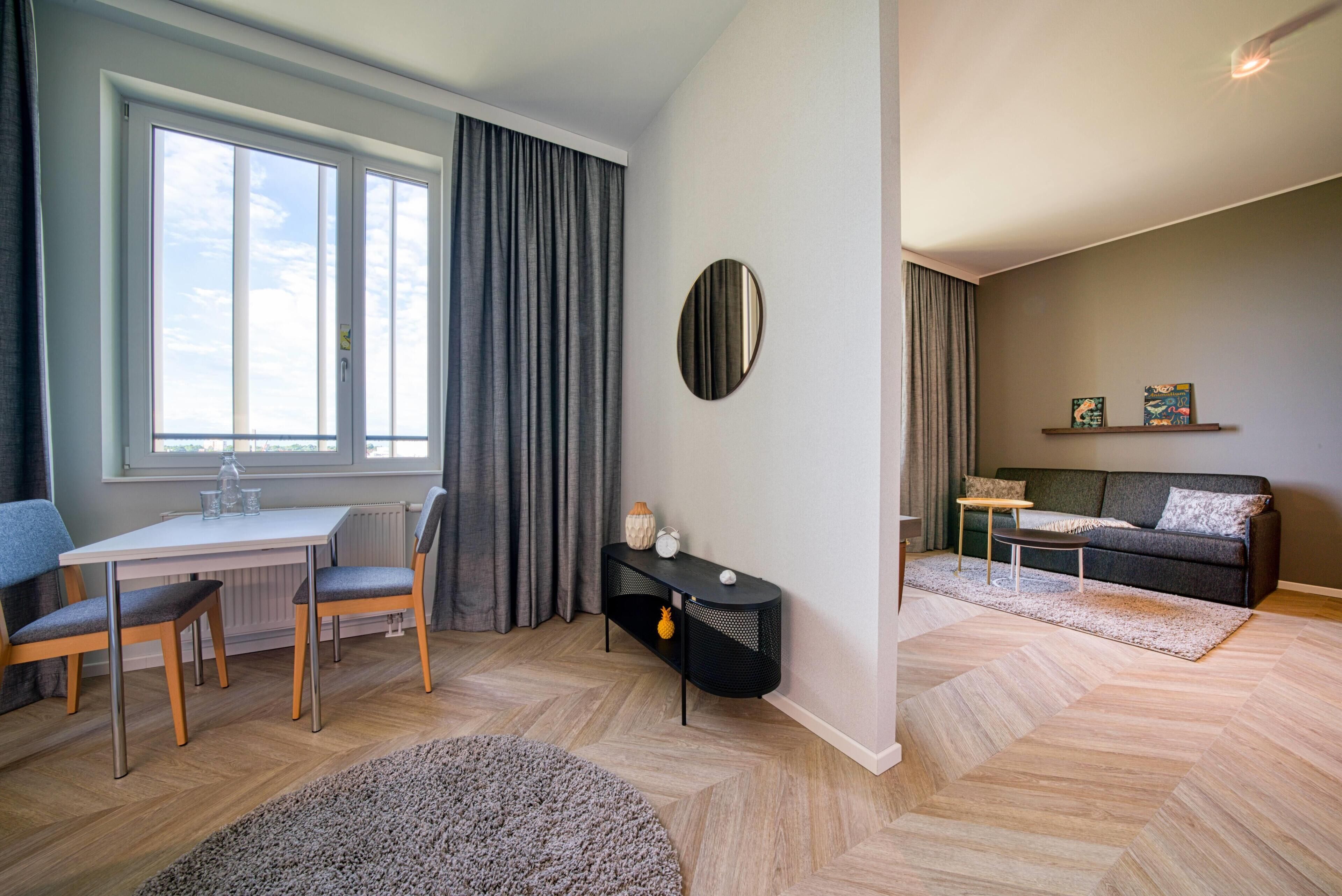 A modern living room with a dining area, featuring a large window, grey curtains, a round mirror, a sofa, a coffee table, and a small side table.