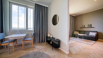 A modern living room with a dining area, featuring a large window, grey curtains, a round mirror, a sofa, a coffee table, and a small side table.