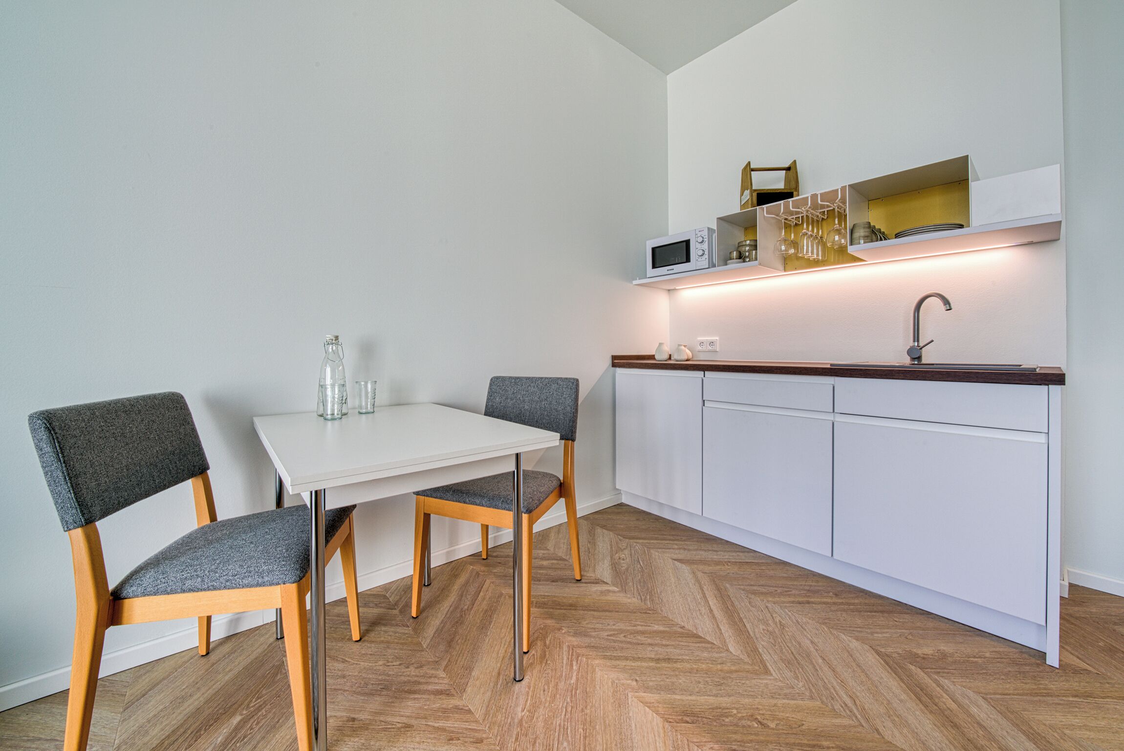 Eine moderne Küche mit weißen Schränken, einem Holzboden im Fischgrätmuster, einem weißen Tisch, zwei grauen Stühlen und einer Mikrowelle auf einem Regal.