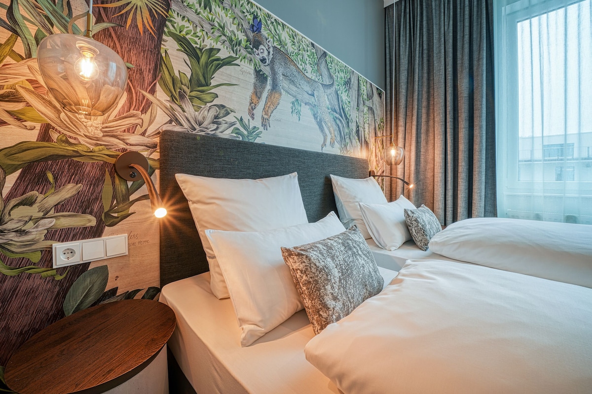 Una habitación de hotel con un mural grande, una cama con sábanas blancas y una mesita de noche con una lámpara.