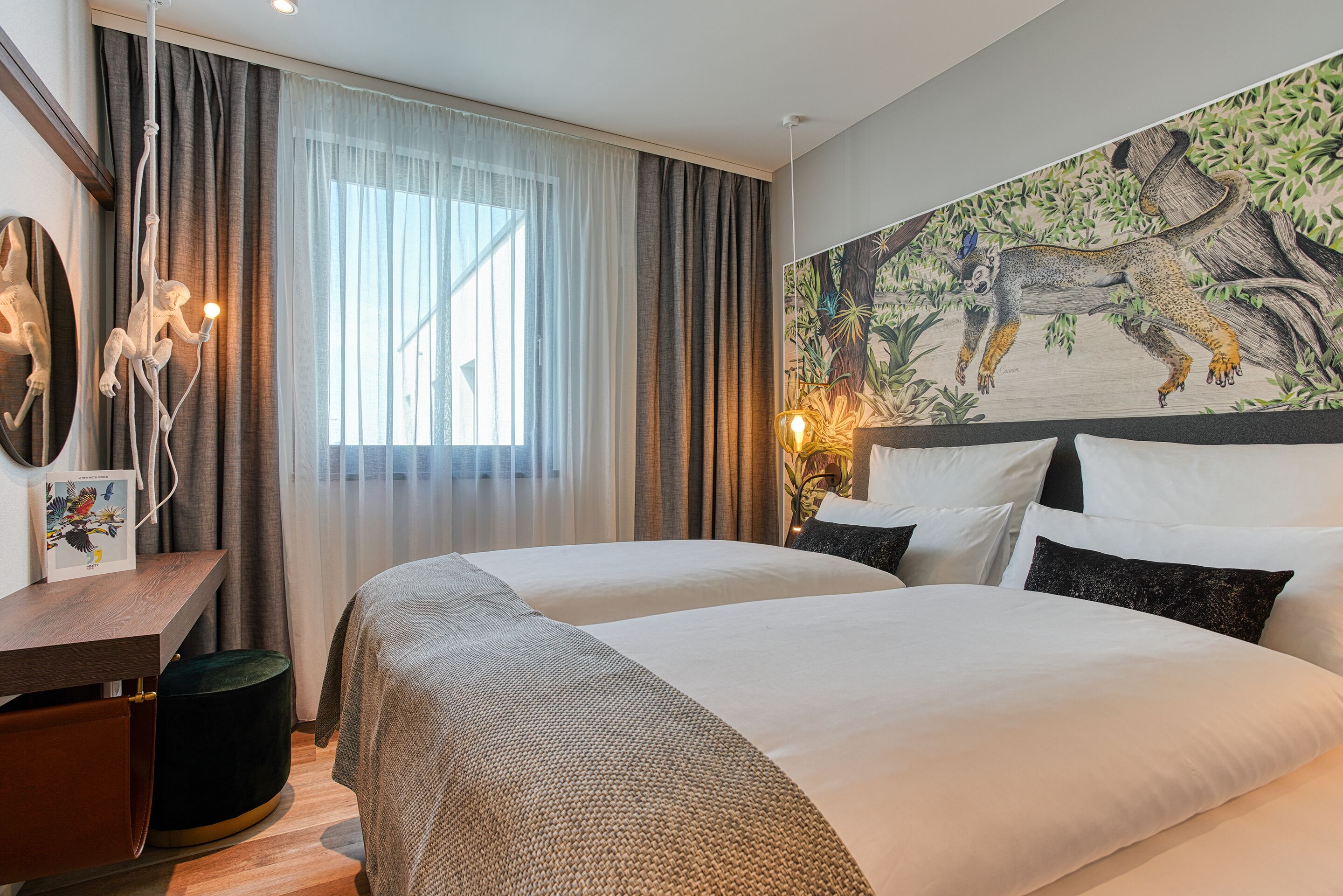 Ein Hotelzimmer mit zwei Betten, einem Schreibtisch mit Stuhl, einem großen Fenster mit Vorhängen und einem Wandgemälde.