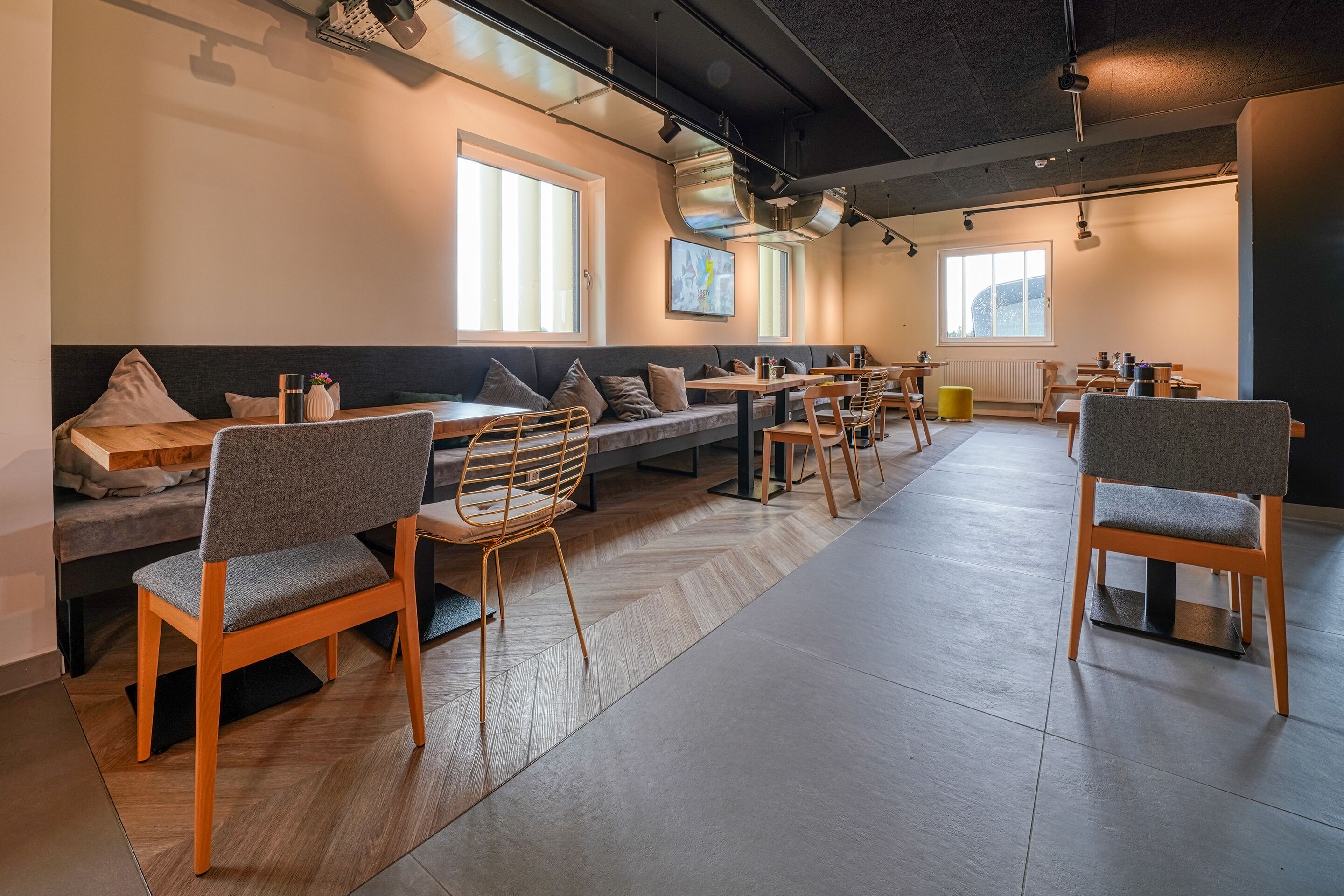A modern dining area with wooden tables and chairs, a long bench, and large windows allowing natural light.