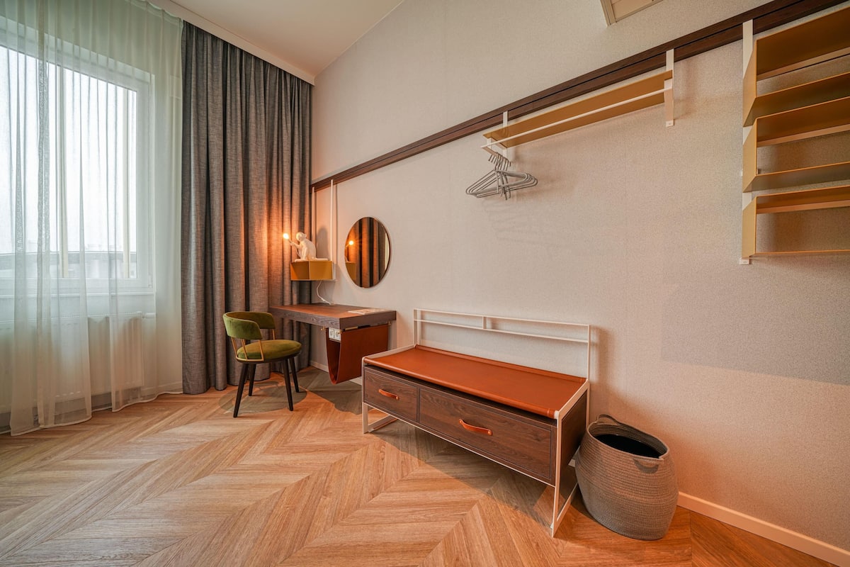 Een hotelkamer met een houten visgraatvloer, een bureau met stoel, een bed met houten hoofdbord, een kledingrek en een groot raam met dunne gordijnen.