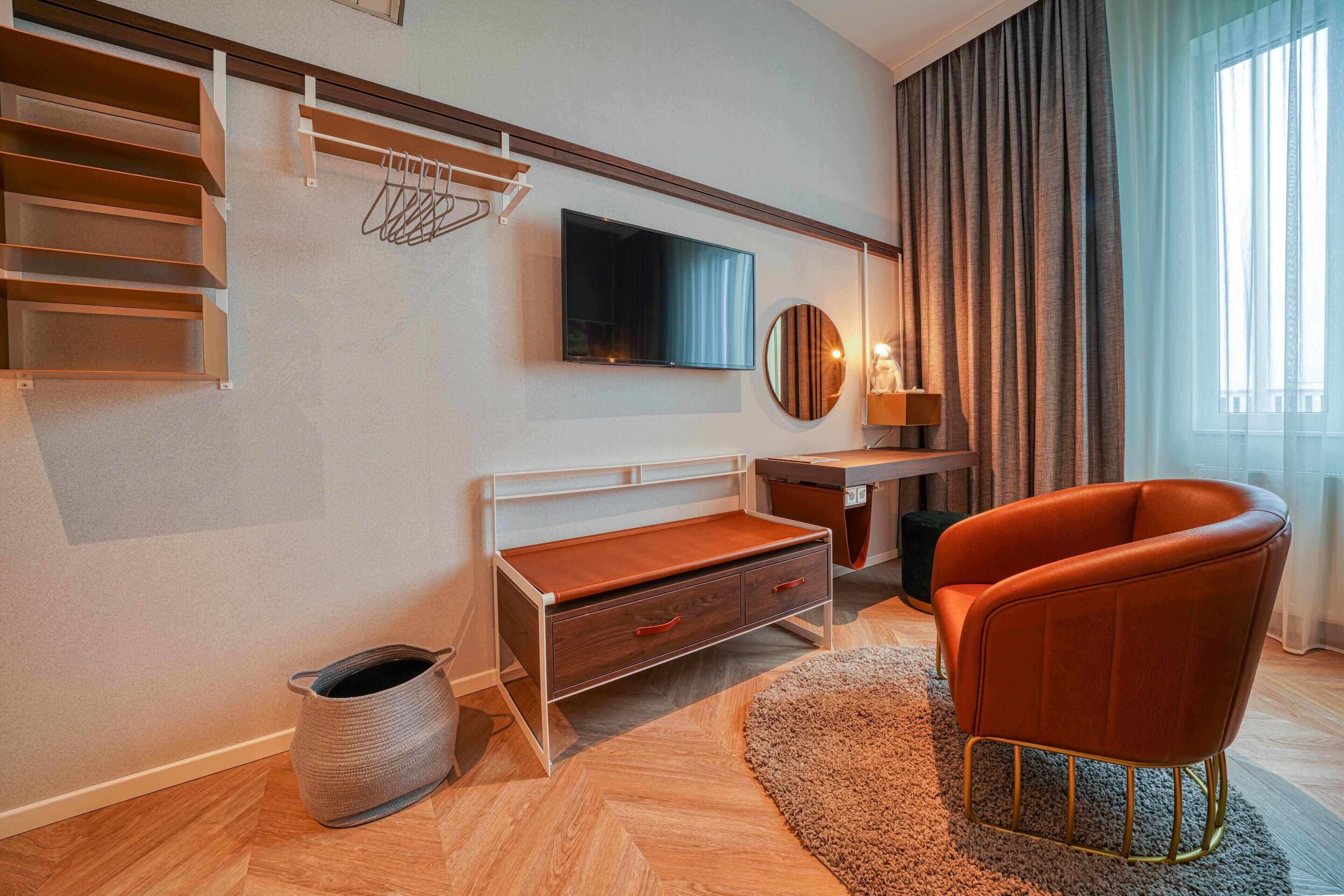 Ein modernes Hotelzimmer mit Holzboden, einem Schreibtisch, einem Flachbildfernseher, einem bequemen orangefarbenen Sessel, einem runden Spiegel und einem großen Fenster mit Vorhängen.