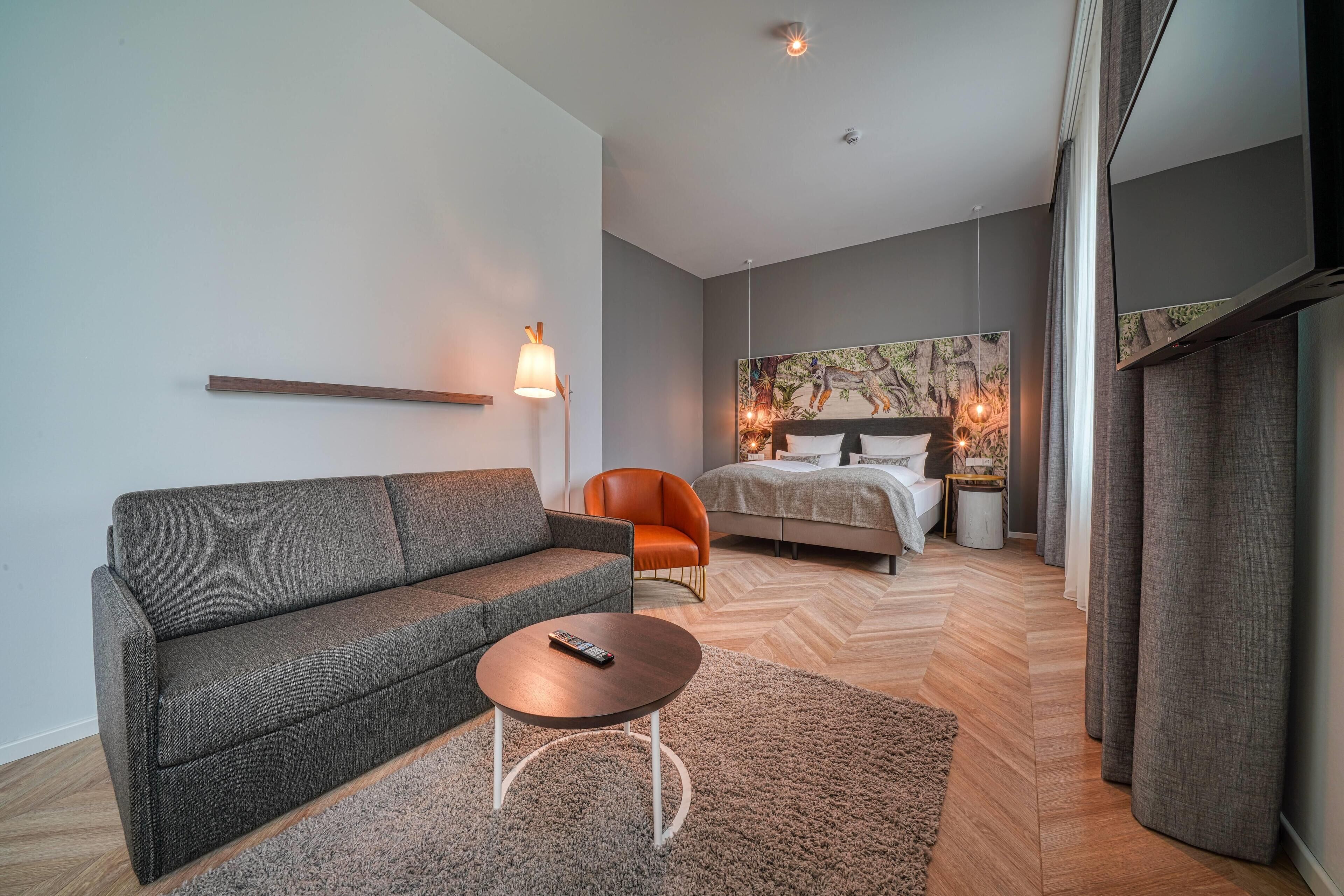 Una habitación de hotel moderna con un sofá gris, una mesa redonda, una butaca naranja, una cama con cabecero y un ventanal grande.