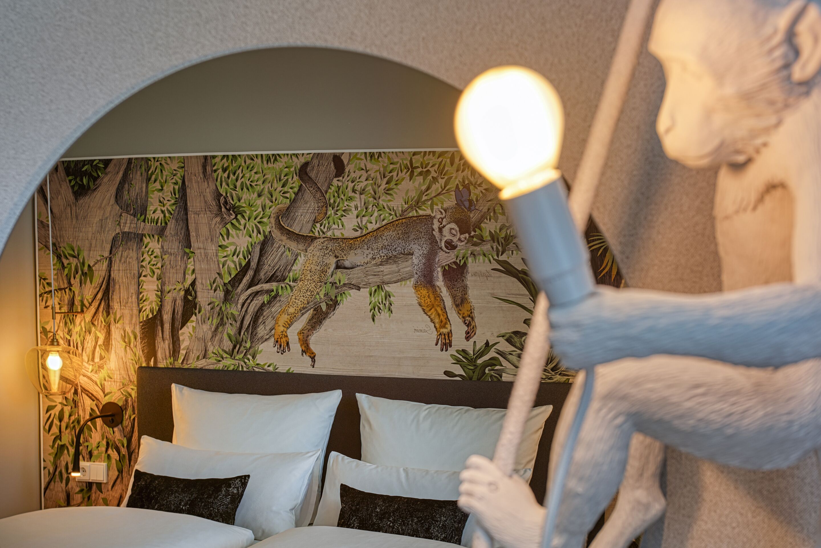 A bedroom with a mural of a deer and tree, a bed with white and dark pillows, and a nightstand with a lamp.
