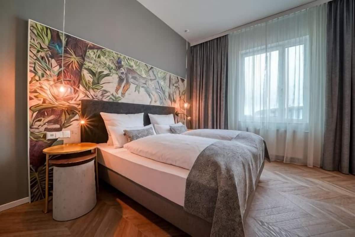 Een slaapkamer met een groot bed, een nachtkastje, een rond bijzettafeltje, een muurschildering en een raam met gordijnen.