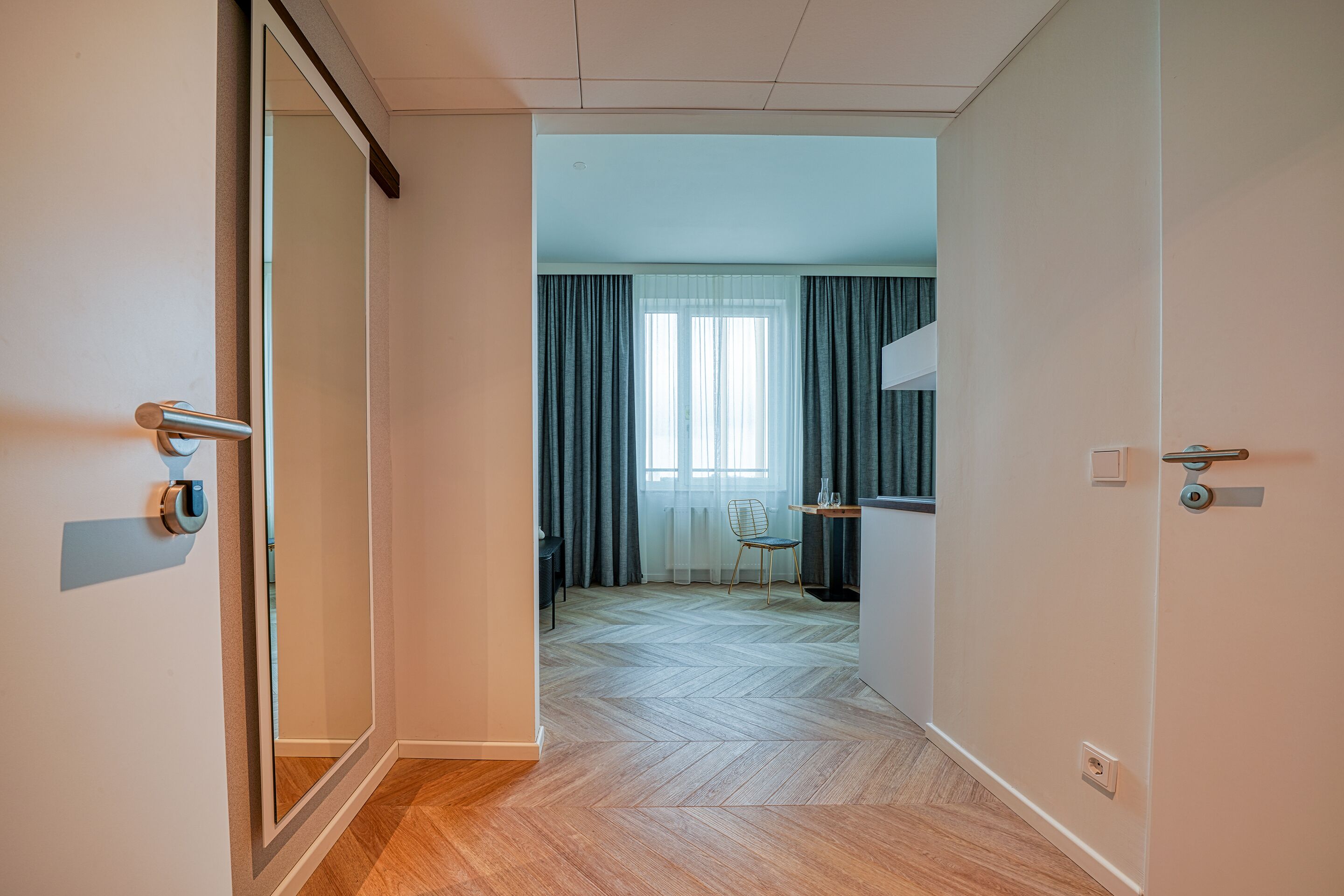 A hallway leading to a room with a herringbone floor, a chair, and a table with a vase.