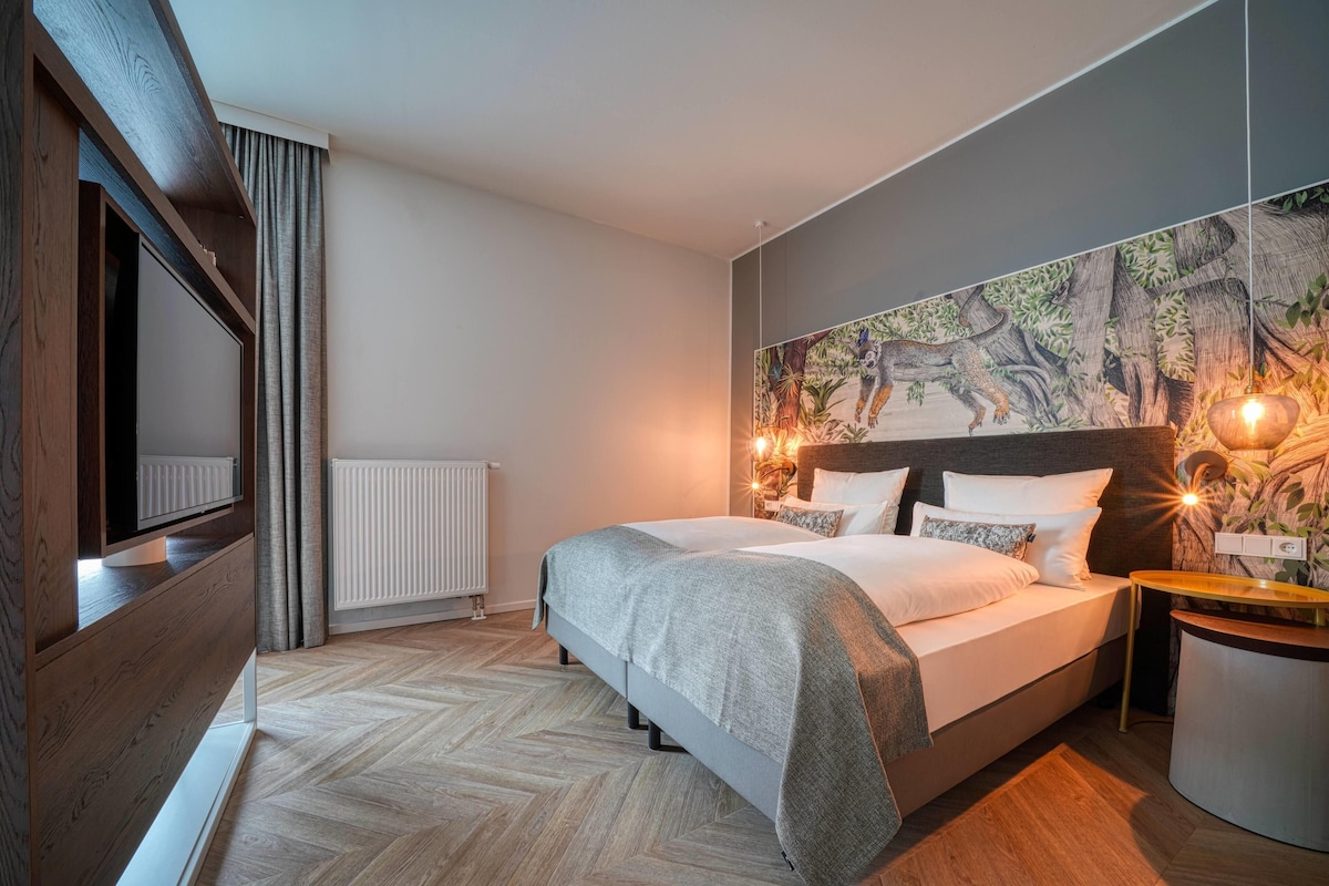 Una habitación de hotel moderna con una cama grande, televisión, un mural en la pared y suelo de madera.