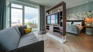 Una habitación de hotel moderna con sofá, televisión, cama y una ventana grande con vistas a edificios.