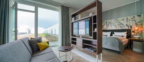 A modern hotel room with a sofa, a TV, a bed, and a large window offering a view of buildings.