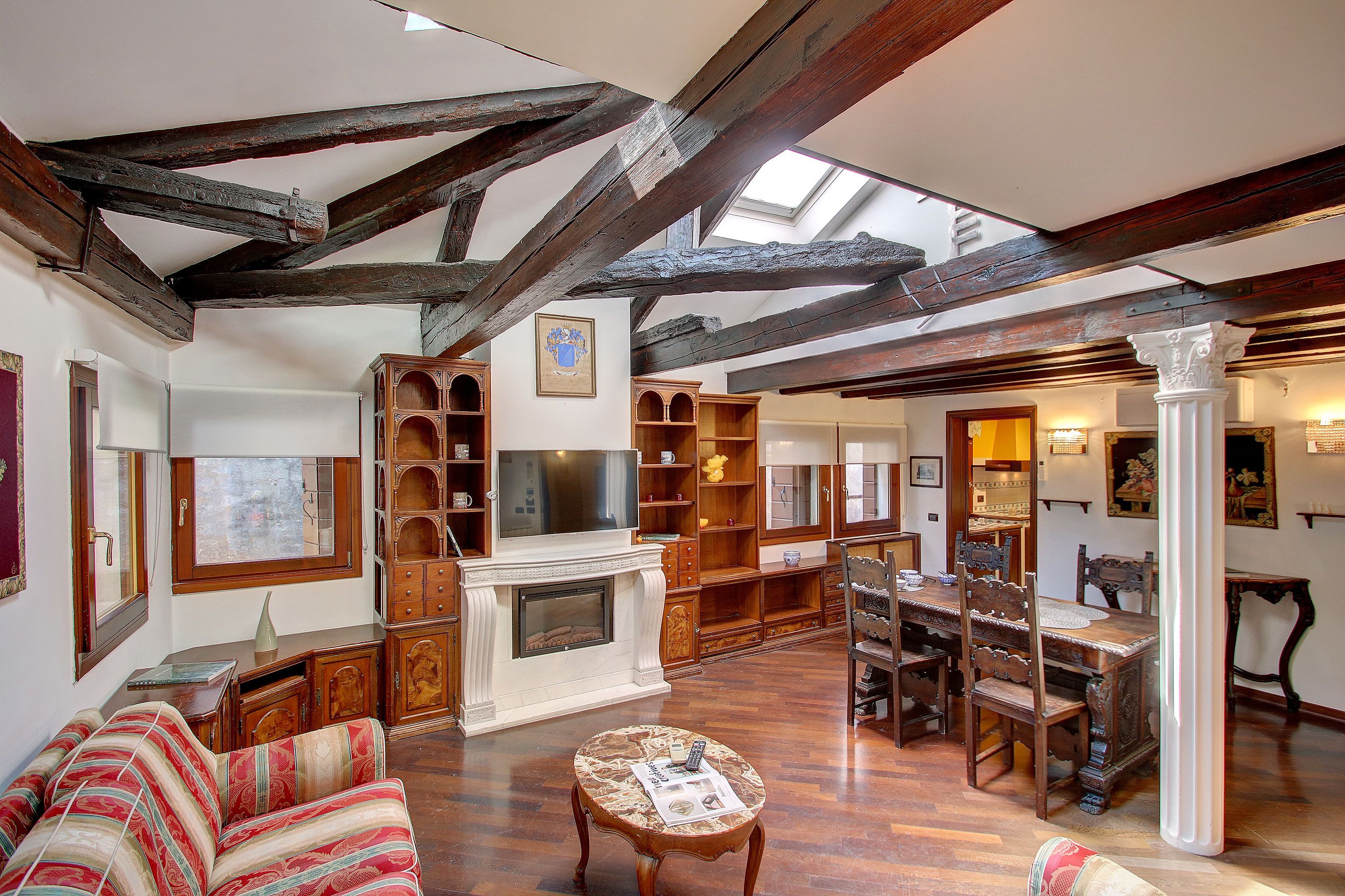A spacious living room with exposed wooden beams, a fireplace, a television, and a dining area with wooden furniture.
