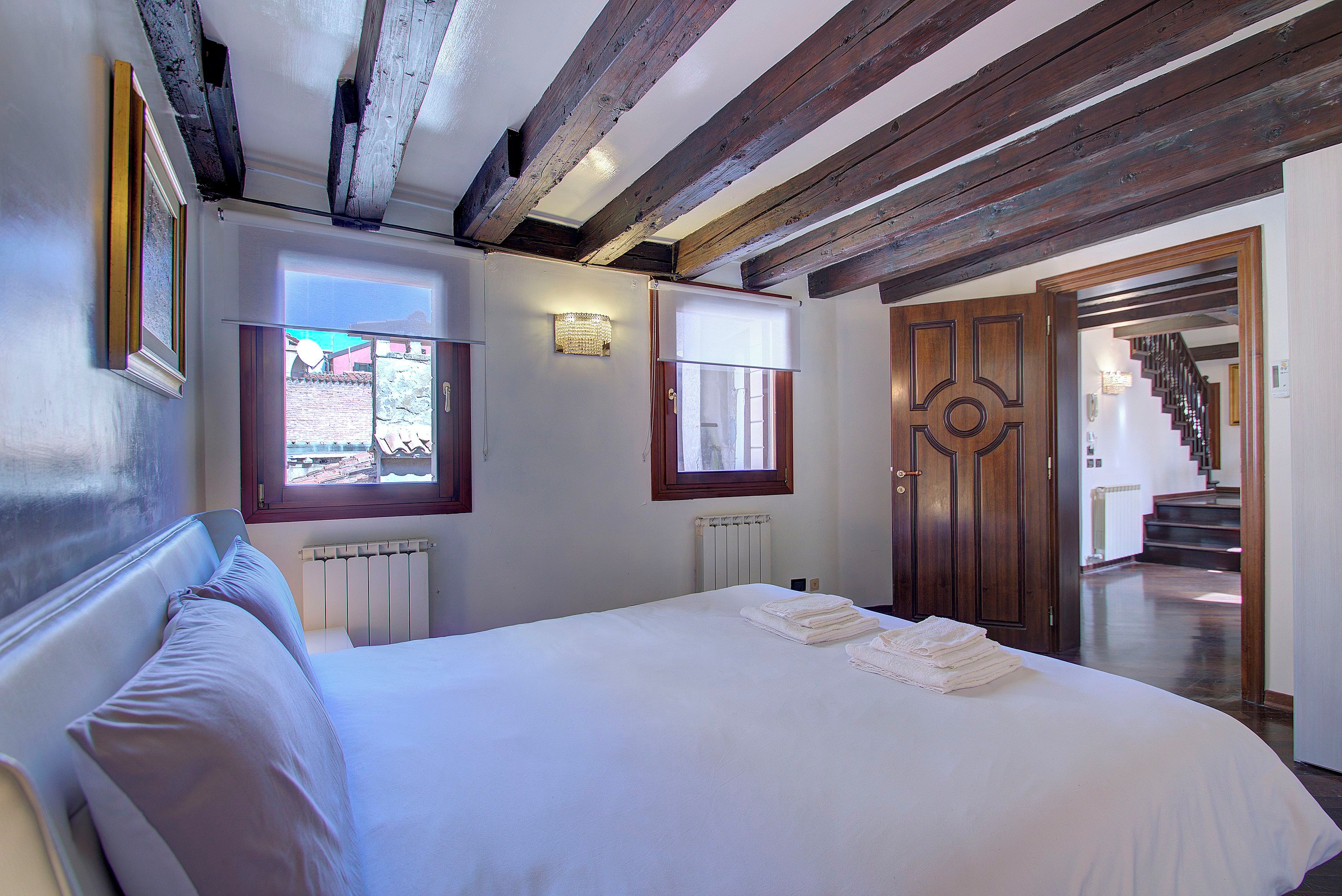 A bedroom with a large bed, wooden beams on the ceiling, a radiator, and a view of the outside.