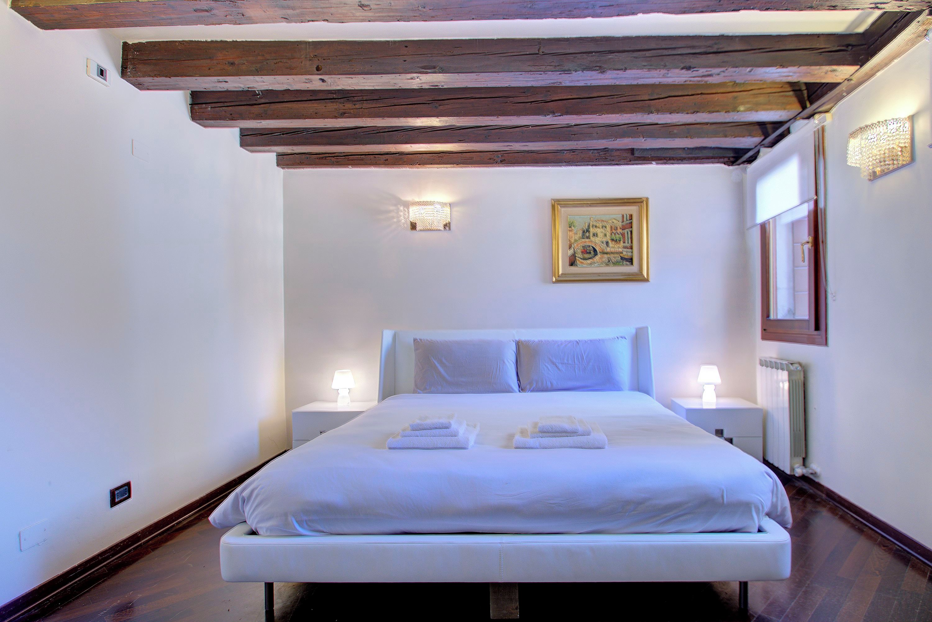 Un lit bien fait, avec du linge de lit et des serviettes blanches, un tableau encadré au mur et deux lampes de chevet.