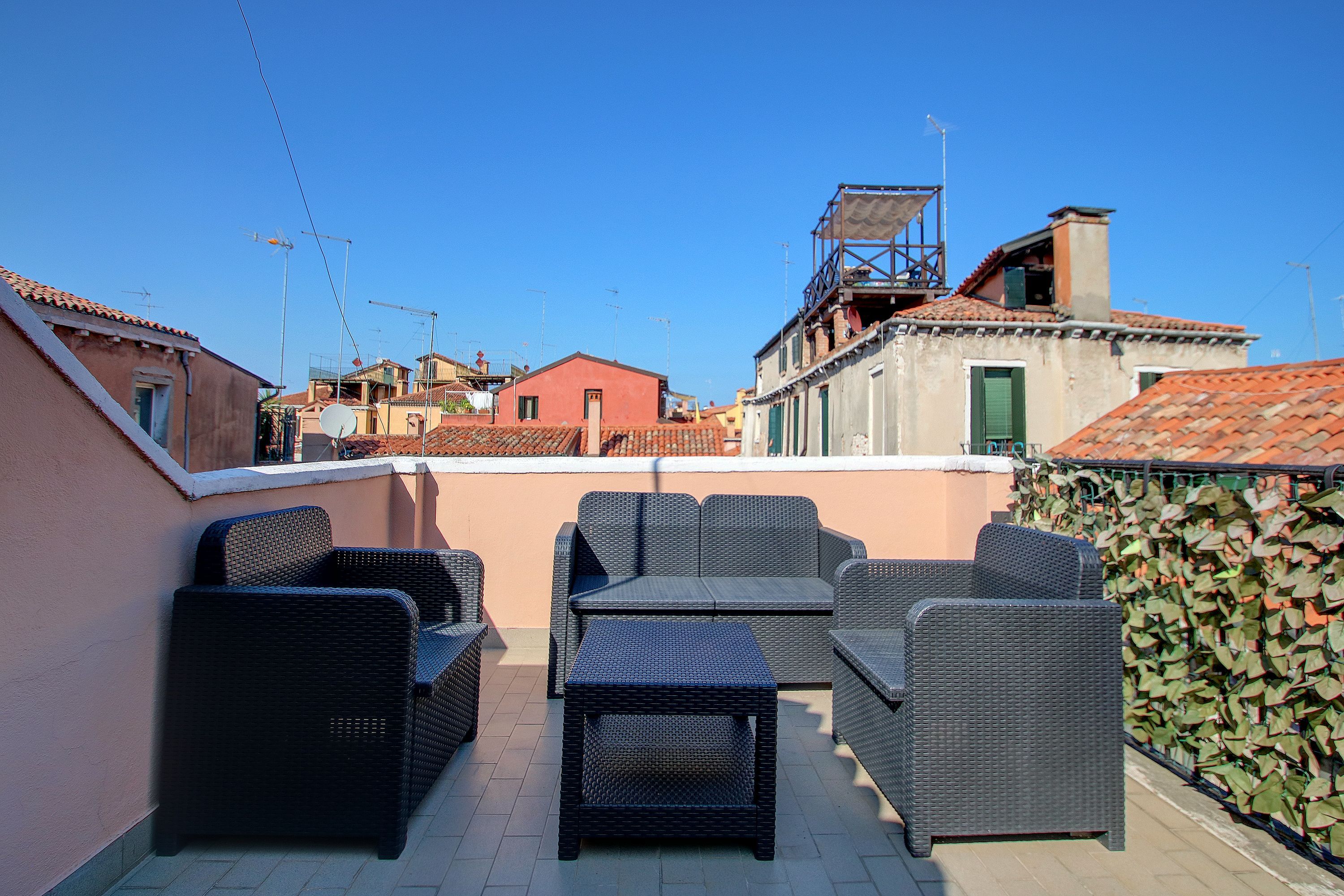 Una terraza en la azotea con mobiliario de mimbre, vistas de edificios circundantes y un cielo azul despejado.