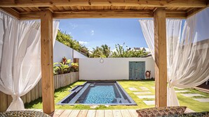 Una terraza de madera con piscina, cortinas blancas y una puerta azul.