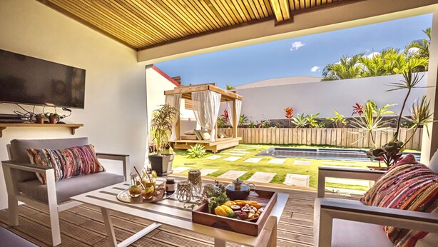 Un área de patio con un sofá, una mesa de centro con una bandeja de frutas, un banco de madera y un televisor montado en la pared.