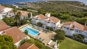 A coastal resort with a swimming pool, lounge chairs, and villas with red-tiled roofs.