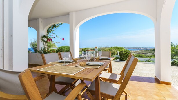 A dining area with a wooden table set for six, surrounded by chairs, with a view of the landscape and sea.