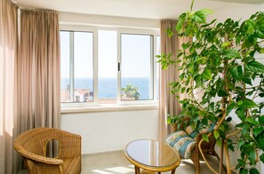 Ein Zimmer mit großem Fenster, einem Korbsessel, einem kleinen runden Tisch und einer Couch.