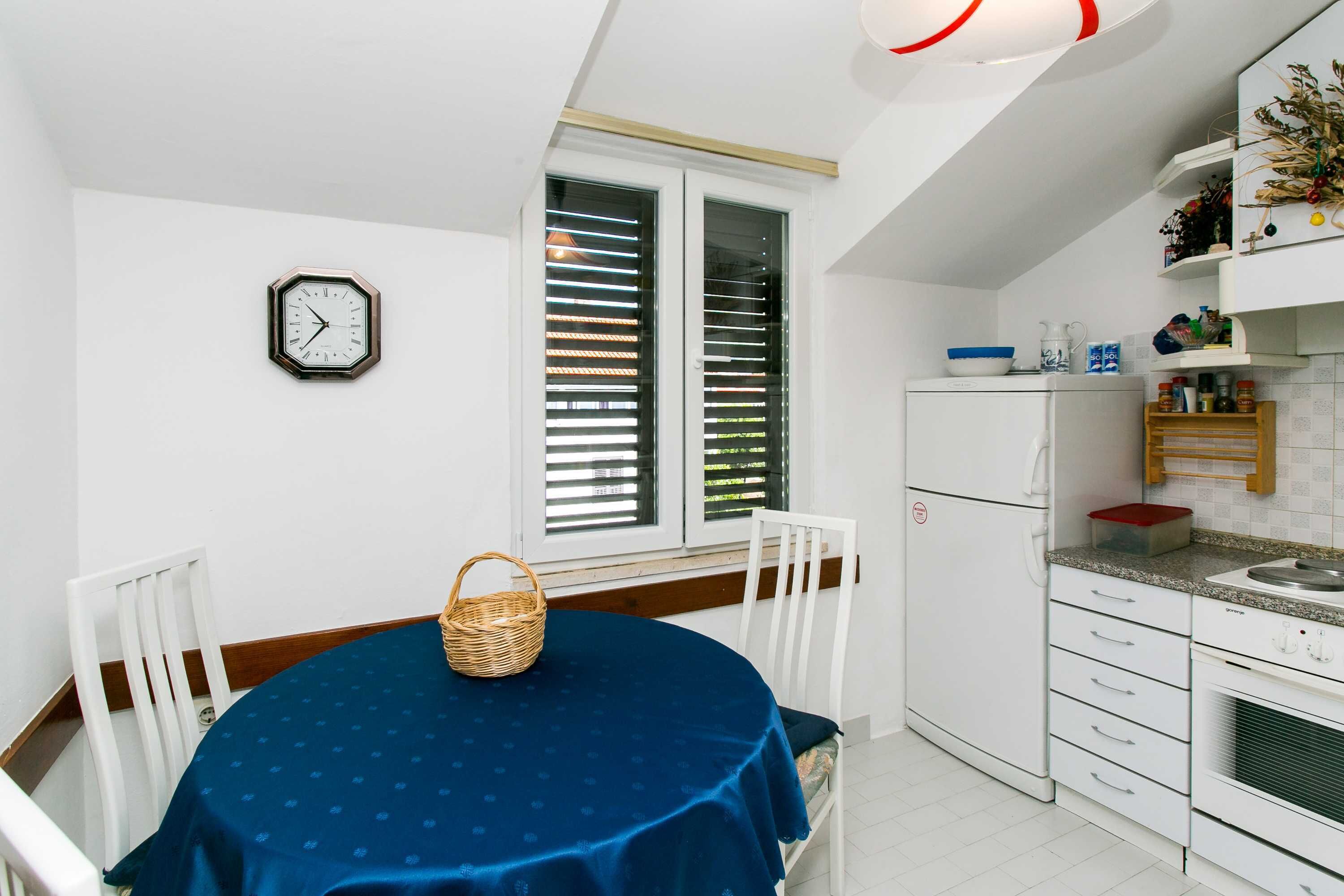 Une cuisine compacte avec des meubles blancs, un réfrigérateur blanc et un coin repas agrémenté d’une nappe bleue.