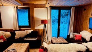 Una habitación acogedora con techo de madera, un gran ventanal con cortinas, un sofá con cojines mullidos, una mesa de madera y una lámpara de pie.
