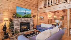 LâintĂ©rieur chaleureux dâun chalet en rondins, avec une cheminĂ©e en pierre, un grand tĂ©lĂ©viseur Ă Ă©cran plat fixĂ© au-dessus, un canapĂ© bleu et un balcon en bois.